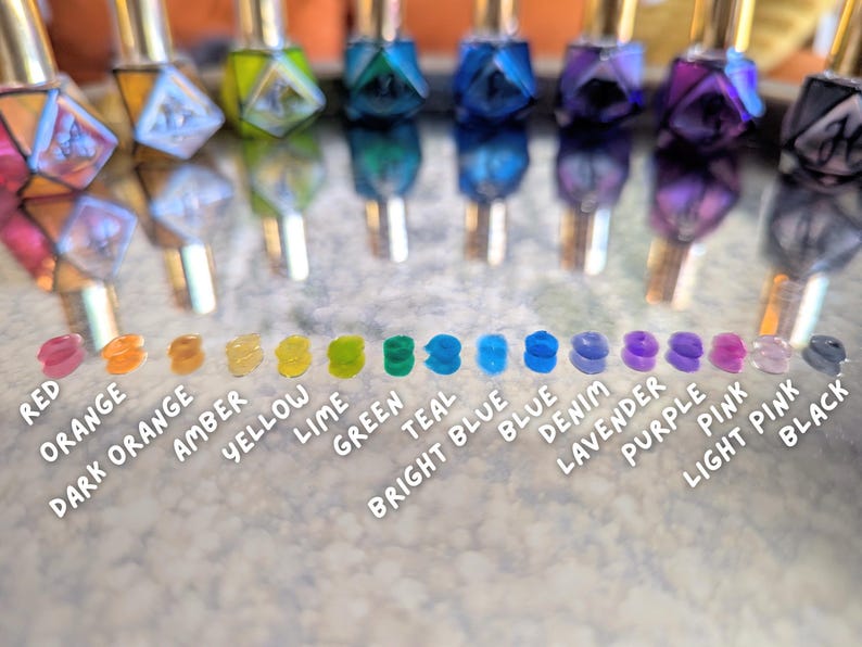 May include: A collection of colorful nail polish bottles with gold caps, reflecting on a surface. Below, a row of matching color swatches labeled with names like "Red," "Orange," "Green," and "Purple."