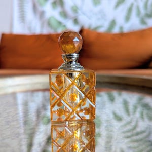 May include: A decorative perfume bottle with a faceted amber-colored stopper and a silver neck. The square bottle has a diamond pattern and is filled with a golden liquid. The bottle is reflected on a mirrored surface.