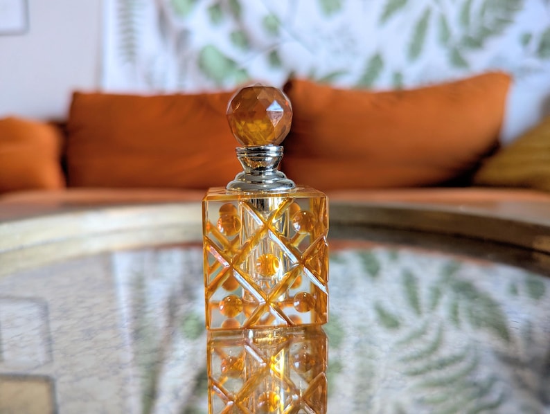 May include: A decorative perfume bottle with a faceted amber-colored stopper and a silver neck. The square bottle has a diamond pattern and is filled with a golden liquid. The bottle is reflected on a mirrored surface.