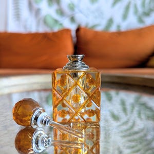 May include: An amber-colored perfume bottle with a silver cap and a decorative stopper. The bottle has a geometric design and sits on a reflective surface. The stopper is lying next to the bottle. The background features orange pillows.