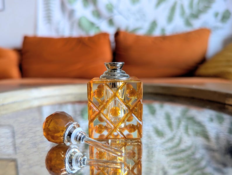 May include: An amber-colored perfume bottle with a silver cap and a decorative stopper. The bottle has a geometric design and sits on a reflective surface. The stopper is lying next to the bottle. The background features orange pillows.