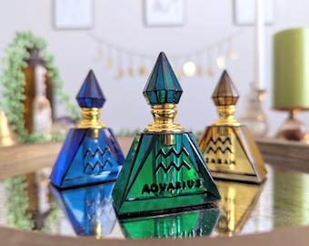 May include: Three pyramid-shaped perfume bottles with gold accents and geometric designs. The bottles are in shades of blue, green, and gold. The word "AQUARIUS" is visible on one bottle.
