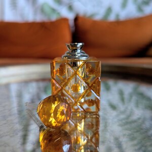 May include: A decorative perfume bottle with a geometric design. The bottle is a warm amber color with a silver cap. The round stopper is next to the bottle. The background is blurred, with orange pillows and a patterned surface.