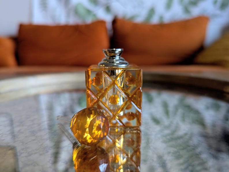 May include: A decorative perfume bottle with a geometric design. The bottle is a warm amber color with a silver cap. The round stopper is next to the bottle. The background is blurred, with orange pillows and a patterned surface.