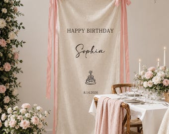 May include: A vertical beige fabric banner hanging from a string with pink ribbon bows at the top corners. The banner features the black text HAPPY BIRTHDAY, the name Sophia in script, a small birthday cake icon, and the date 8.14.2026.