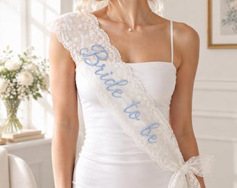 May include: A white lace sash worn diagonally across the torso, featuring the text Bride to be embroidered in light blue script. The sash is made of delicate floral lace with scalloped edges and is tied at the hip with a matching lace ribbon.