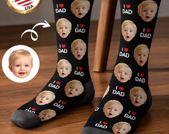 May include: Black socks with a repeating pattern of a baby's face, the words "I ❤️ DAD", and a red heart. The socks have gray toes and heels. A "Made in USA" emblem is in the upper left corner.