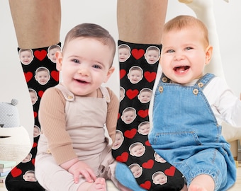May include: Two pairs of black socks with red hearts and a pattern of a baby's face. The socks are worn by two babies, one sitting on the floor and the other standing.