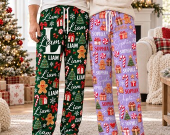 May include: Two pairs of pajama pants with white drawstring waistbands. The left pair is dark green with a repeating pattern of gingerbread men, Christmas trees, gift boxes, candy canes, and the name Liam. The right pair is light purple with the same pattern and the name Sophia.