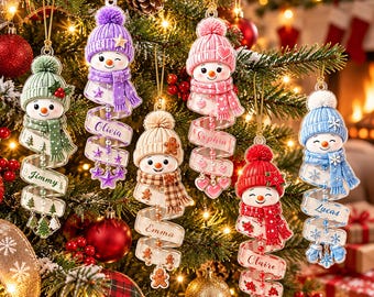 May include: Six snowman ornaments hanging on a Christmas tree. Each snowman wears a colored knit hat and scarf, with a spiraling ribbon below featuring names like Jimmy, Olivia, Emma, Sophia, Claire, and Lucas, plus festive charms.