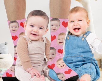 May include: Two pairs of pink socks with red hearts and a pattern of baby faces. The socks are worn by two babies, one sitting and one standing.