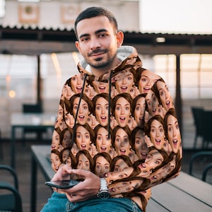 May include: A black hooded sweatshirt with a repeating pattern of a person's face. The face is smiling and has a surprised expression.