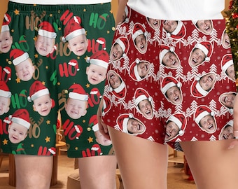 May include: Green and red Christmas-themed shorts. The green shorts feature baby faces wearing Santa hats, the word "HO HO HO", and gold stars. The red shorts have a pattern of laughing faces wearing Santa hats and white Christmas trees.