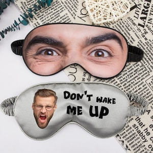 May include: Two gray sleep masks. One has a man's face printed on it with the words "Don't Wake Me Up". The other has a man's face with his eyes wide open.