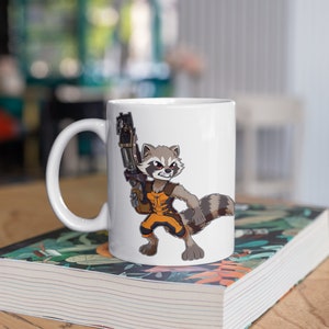 Może przedstawiać: Biały ceramiczny kubek z postacią z kreskówki szopa w pomarańczowo-czarnym stroju, trzymającą dużą broń. Kubek stoi na książce z kwiatowym wzorem, z rozmytym tłem.