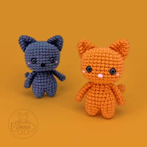 May include: Two handmade crochet cat dolls. One is dark blue, the other is orange. Both have black eyes, pink cheeks, and small arms and legs. The dolls are standing on an orange background. The dolls are approximately 4 inches tall.