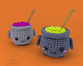 Op de afbeelding: Twee gehaakte ketel amigurumi speelgoed met lachende gezichten. De ene ketel is gevuld met paarse drank en de andere met groene drank. Beide ketels hebben een houten roerstaaf.