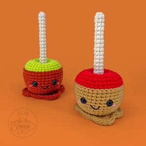 Puede incluir: Dos manzanas de caramelo de crochet con caras sonrientes. Una manzana de caramelo es roja y verde, y la otra es roja y marrón. Ambas manzanas de caramelo tienen palos blancos.