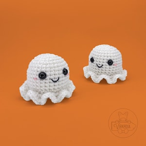 May include: Two handmade, white crochet ghosts with black eyes, pink cheeks, and smiling mouths. The ghosts have a ruffled bottom edge and are set against an orange background. These cute, whimsical ghosts are perfect for Halloween decorations.