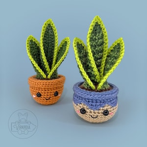 May include: Two crocheted snake plants in decorative pots. The plants have green and yellow leaves. One pot is orange, the other is blue and tan. Both pots have smiling faces and are set against a blue background.
