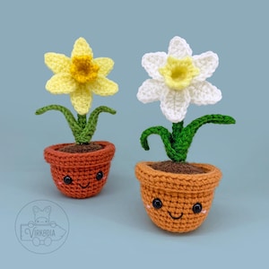 Peut inclure: Deux plantes de jonquilles crochetées dans des pots en terre cuite. L'une est jaune avec un centre jaune, l'autre est blanche avec un centre jaune. Les pots ont des visages souriants sur un fond bleu.