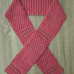 May include: A handmade, dusty rose-colored crochet scarf. The scarf features a textured, woven pattern with openwork detailing near the ends. The scarf is displayed on a wooden surface.