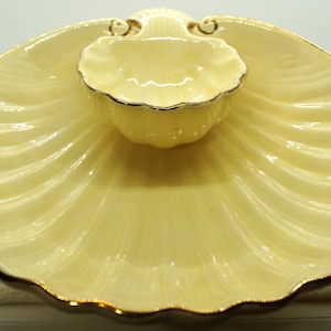 May include: A yellow ceramic serving dish shaped like a seashell with a smaller bowl in the center. The dish and bowl have gold trim.