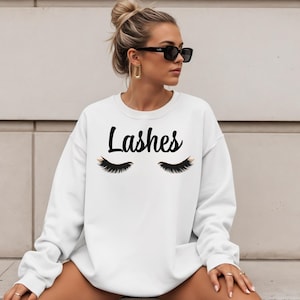 May include: White sweatshirt with the word "Lashes" in black script above a graphic of closed eyelashes. The eyelashes are black with gold accents. The sweatshirt is a crew neck style.
