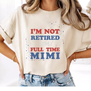 May include: A cream-coloured short-sleeve t-shirt featuring the red and blue text I'M NOT RETIRED I'M FULL TIME MIMI. The design includes small red and blue star accents and scattered dots, creating a patriotic-themed graphic on the front chest.