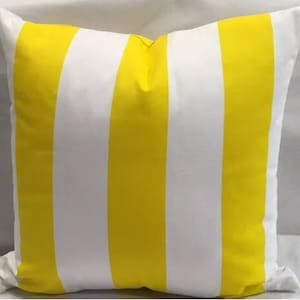 Puede incluir: Cojín decorativo cuadrado con un llamativo patrón de rayas verticales amarillas y blancas. La almohada tiene un aspecto suave y texturizado, probablemente de tela. El diseño es simple y moderno, adecuado para varios estilos de decoración del hogar.