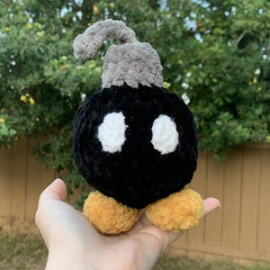 May include: A handmade crochet Bob-omb plush toy. The plush is black with white eye details and yellow feet. It has a gray fuse and top. The toy is held in a hand.
