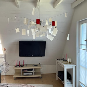 Könnte beinhalten: Ein moderner Kronleuchter im Sputnik-Stil mit dünnen Metallarmen, die von einer zentralen Nabe ausgehen. Die Leuchte hält zahlreiche rechteckige weiße Papierkarten und zwei rote Papierkarten, die mit kleinen Clips an den Enden befestigt sind.