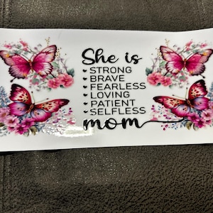May include: A white background with pink flowers and butterflies. The text "She is" is written in black, followed by a list of qualities: strong, brave, fearless, loving, patient, selfless. The text "mom" is written in black below the list.