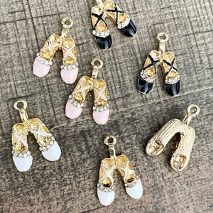 Puede incluir: Un conjunto de seis dijes de bailarina de color dorado. Los dijes son de tres colores diferentes: negro, blanco y rosa. Cada dije presenta un par de zapatos de bailarina con una cinta dorada y diamantes de imitación.