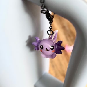 Könnte beinhalten: Ein lila Axolotl-Anhänger mit einem süßen Gesicht und schwarzen Augen hängt an einer schwarzen Kette. Der Anhänger ist aus Polymer Clay gefertigt und hat eine glänzende Oberfläche.