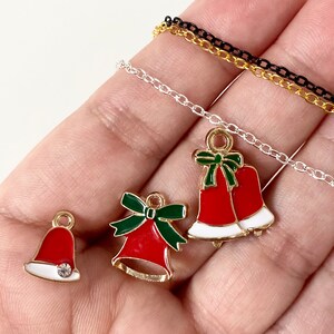 Pode incluir: Três pingentes de sino de Natal dourados com esmalte vermelho e verde e uma base branca. Um pingente tem um laço verde, outro tem um pompom branco e outro tem dois sinos. Os pingentes estão em uma corrente prateada e uma corrente preta.