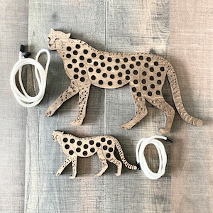May include: Two wooden cheetah shapes with holes for lacing. The larger cheetah is brown with black spots and the smaller cheetah is brown with black spots. Two white cords are included for lacing.