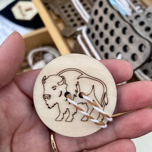 Puede incluir: Un botón redondo de madera clara con un contorno detallado de un bisonte. El botón tiene dos agujeros con hilo marrón y blanco para coser. El botón se sostiene en una mano.