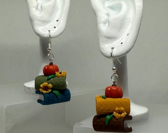May include: A pair of dangle earrings featuring stacks of miniature books in blue, green, and brown, topped with a small orange pumpkin and a yellow sunflower. The earrings hang from silver-colored hooks.