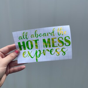 以下が含まれることがあります： 転写紙の白いシートに、「all aboard the hot mess express」と書かれた緑と金のビニールステッカーが貼られています。