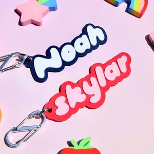 May include: Two personalized keychains with the names "Noah" and "Skylar" in white lettering. The "Noah" keychain has a navy blue border, while the "Skylar" keychain has a red border. The keychains are attached to silver carabiner clips. Scattered around are colorful decorative shapes.