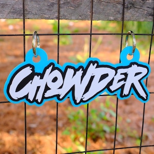 May include: A blue keychain with the word "CHOWDER" in white, outlined in black, and set against a blue background. The keychain is hanging on a black wire fence.