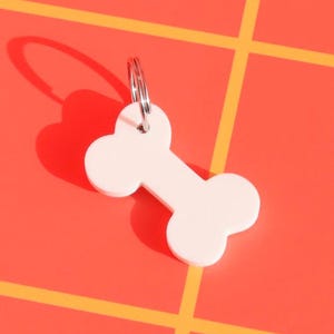 May include: White bone-shaped keychain with a silver ring. The keychain is on a red and yellow checkered background.