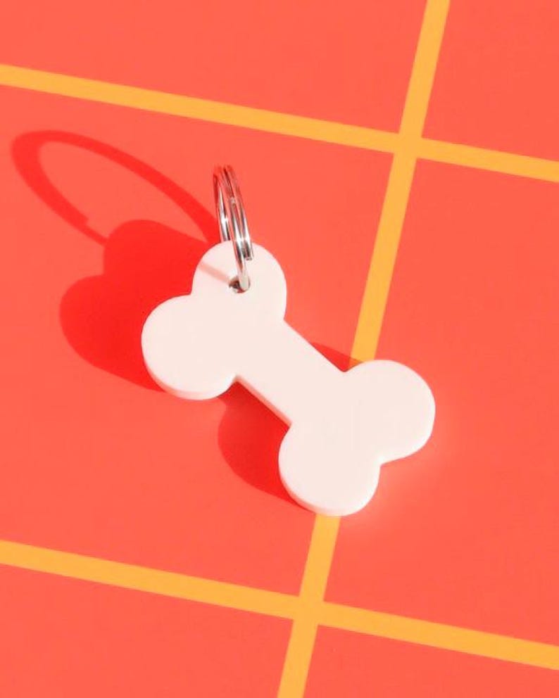 May include: White bone-shaped keychain with a silver ring. The keychain is on a red and yellow checkered background.