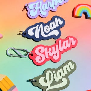 May include: Colorful name keychains in purple, navy, red, and green with white lettering. The names "Harper", "Noah", "Skylar", and "Liam" are displayed. A rainbow and pencil-shaped charm are also visible. The keychains have silver-colored clasps.