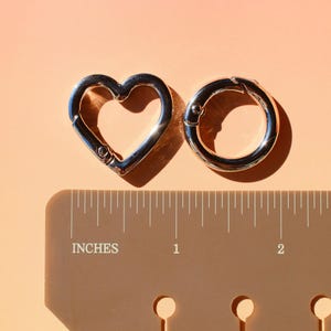 May include: Two silver-colored metal clasps, one heart-shaped and one circular, are displayed on a peach-colored surface. A ruler with inch measurements is placed below the clasps. The heart clasp is approximately 1 inch wide.