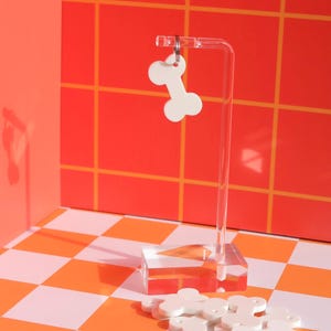 May include: White bone-shaped dog tags on a clear acrylic stand against a red and white checkered background. The tags are arranged in a pile and have a silver ring attached to each one.