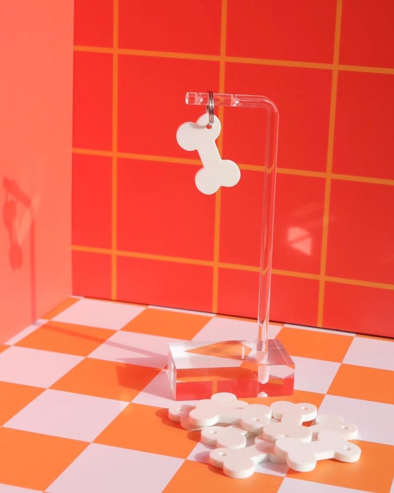 May include: White bone-shaped dog tags on a clear acrylic stand against a red and white checkered background. The tags are arranged in a pile and have a silver ring attached to each one.