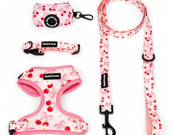 May include: A pet accessory set featuring a pink harness, collar, leash, and waste bag dispenser, all decorated with a cherry and bow pattern. Each item includes a black label with the white text Duck & Luca and black hardware.