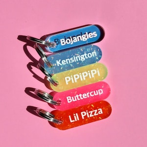 May include: Five colorful keychains with glitter embedded in the plastic. The keychains are labeled "Bojangles", "Kensington", "PiPiPiPi", "Buttercup", and "Lil Pizza".